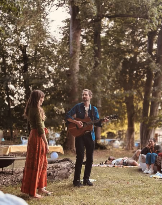 Two musicians performing outdoors among trees as listeners sit on the grass